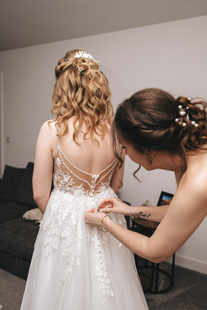 A woman in a white lace wedding dress embellished with intricate beading and a sheer back stands as another woman helps button her gown at Hall Farm Hotel. The bride's wavy hair is adorned with floral accessories. They are indoors, with a couch and a small framed photo partially visible in the background. © Aimee Lince Photography