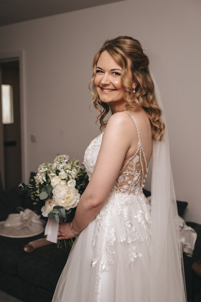 A bride with curly blonde hair smiles, holding a bouquet of white and green flowers. She wears a white lace wedding dress with intricate back detailing and a veil. At the Hall Farm Hotel, the room has neutral-colored walls, and there's a couch in the background with a white cloth draped over it. © Aimee Lince Photography