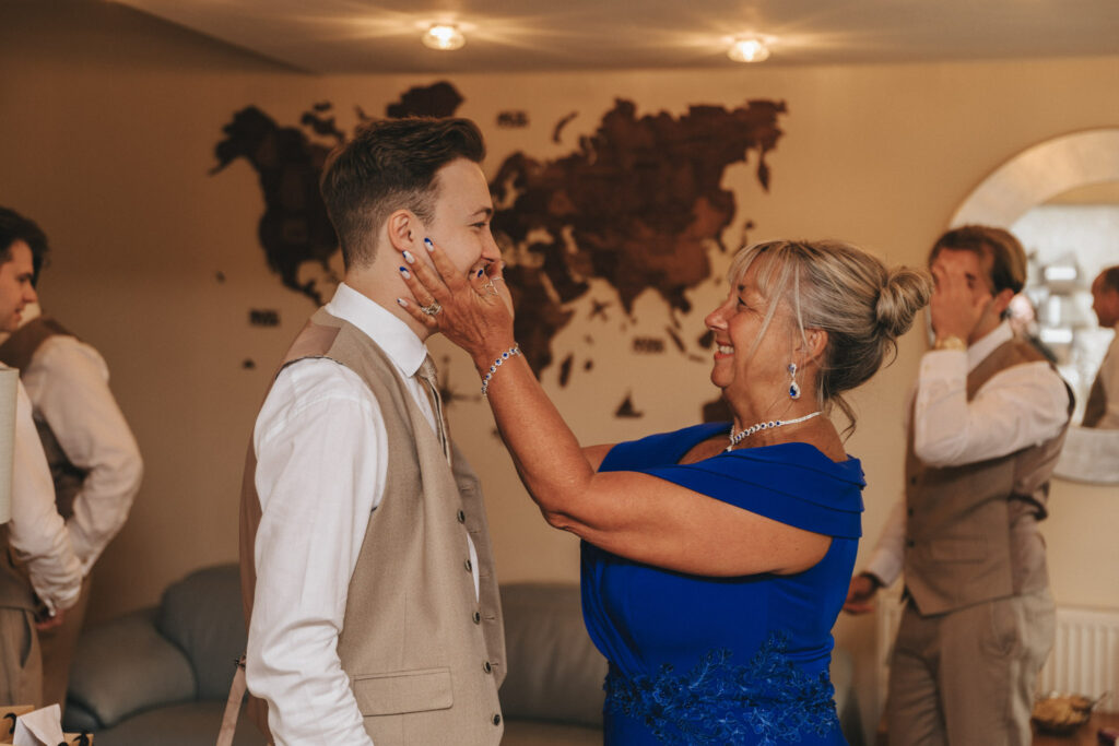 Hall Farm Hotel Wedding Photographer - Courtney & Tom © Aimee Lince Photography (95) A bride with long, wavy blonde hair shares a tender mother-daughter moment on her wedding day, facing an older woman in a floral dress and teal hat. The older woman smiles warmly near a cream-colored wardrobe, as the room's vintage wallpaper adds to this cherished scene. © Aimee Lince Photography - Wedding photographer in Lincolnshire, Yorkshire & Nottinghamshire