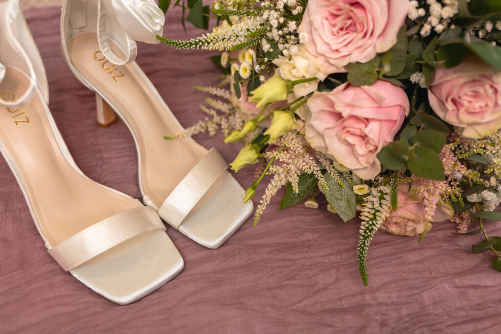 A close-up of elegant white open-toe high heels with satin ankle bows, placed on a mauve textured surface at Hall Farm Hotel. Next to the shoes, a bouquet of pink roses, white flowers, and greenery creates a soft, romantic atmosphere reminiscent of Waltham's charming allure. © Aimee Lince Photography