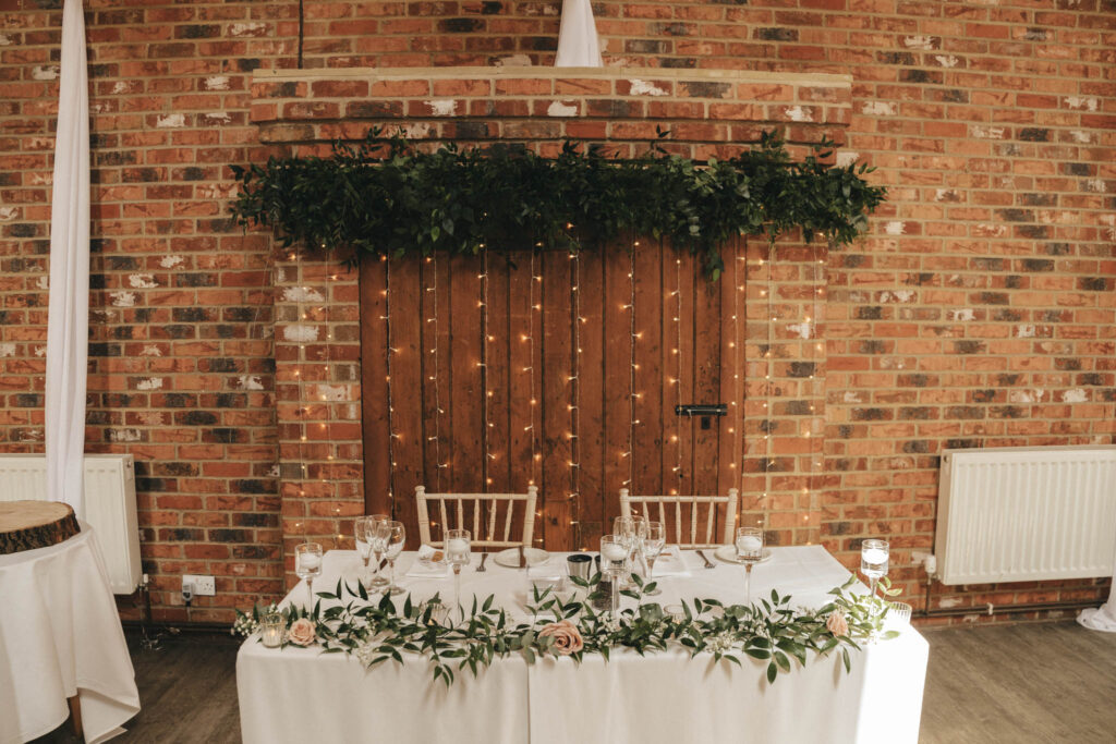 A rustic wedding setup at Hall Farm Hotel in Lincolnshire features a brick wall with a wooden backdrop adorned with greenery and string lights. In front, a table is set with elegant glassware, floral arrangements, and candles. Two white chairs are positioned behind the table, creating a romantic atmosphere. © Aimee Lince Photography
