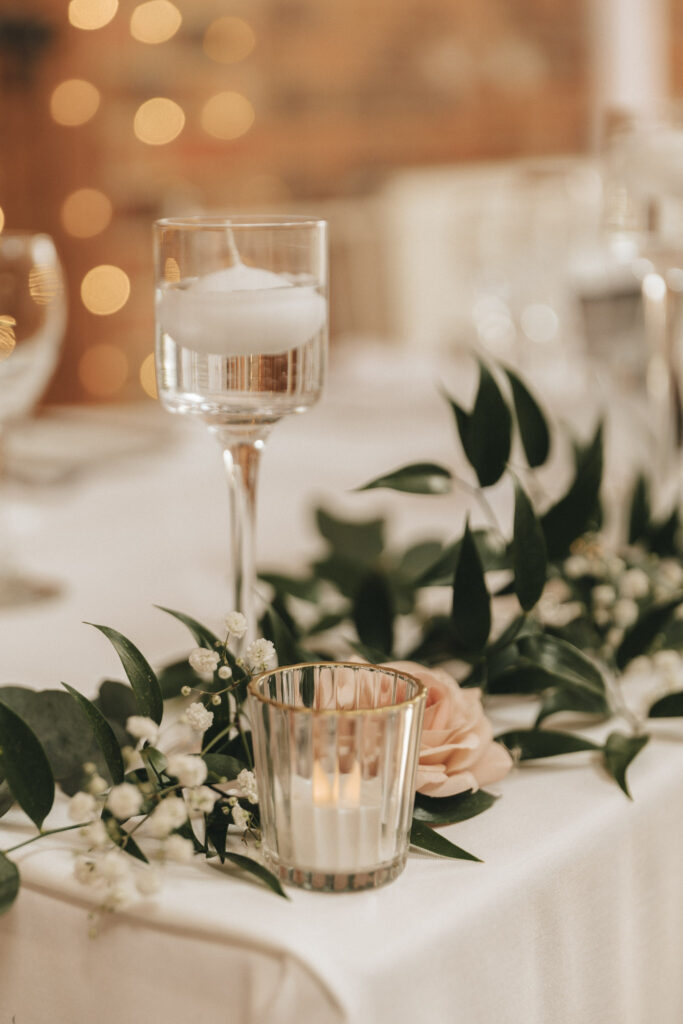 A beautifully set table at the Hall Farm Hotel features a tall, clear candleholder with a floating candle beside a metallic votive. Greenery and small white flowers adorn the table, with a hint of peach rose, creating an elegant and romantic atmosphere. Blurred lights enhance this Grimsby setting. © Aimee Lince Photography