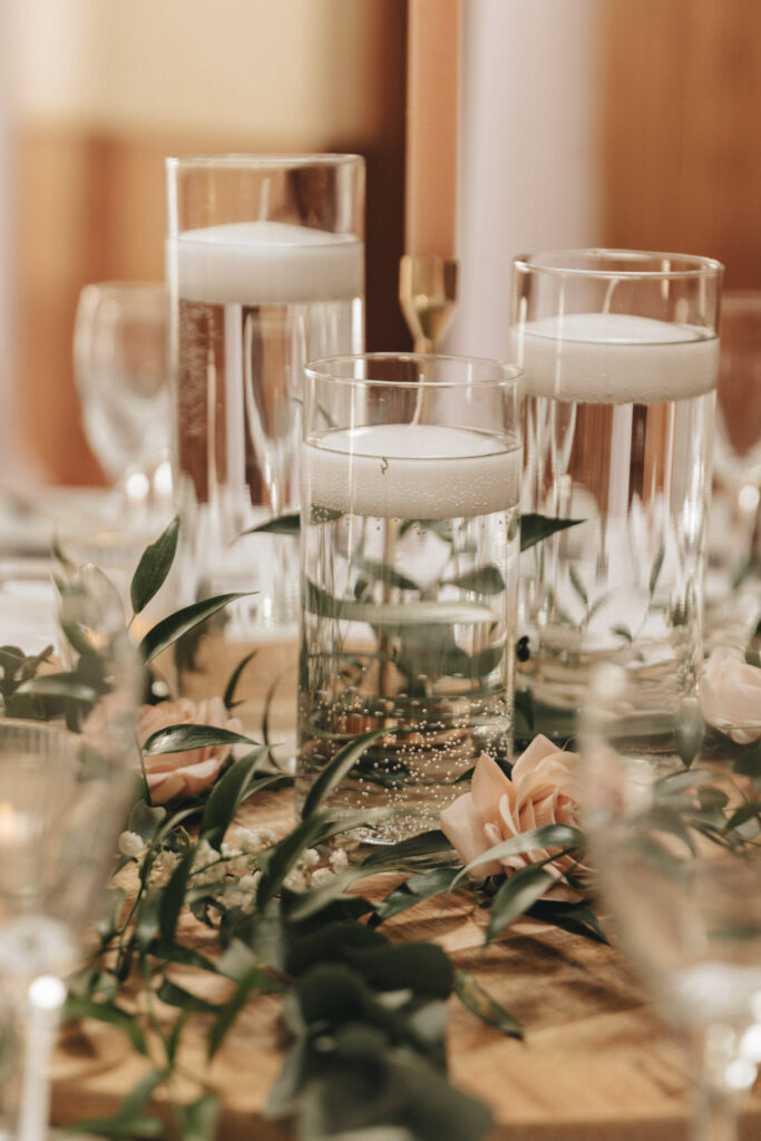 At the exquisite Hall Farm Hotel in Grimsby, an elegant table setting features three cylindrical glass vases filled with water and floating candles. Greenery and blush roses are artfully arranged around the vases, while blurred wine glasses and a tall, copper candle holder add sophisticated charm. © Aimee Lince Photography