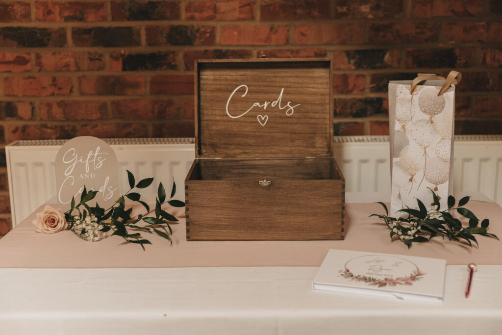 A wooden card box labeled "Cards" with a heart symbol sits on a table at the Grimsby wedding. Next to it is a decorative gift bag with a balloon pattern and greenery. A round sign reads "Gifts and Cards," and a floral guestbook completes the charming setup against the brick wall in Lincolnshire. © Aimee Lince Photography