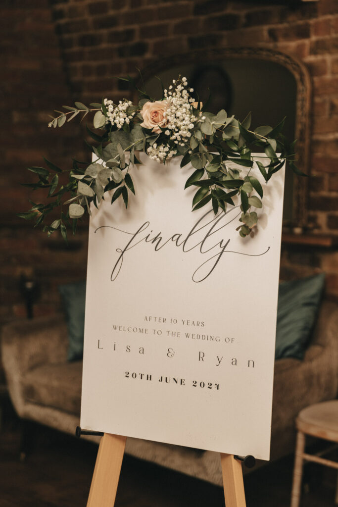 A wedding sign on an easel reads: "Finally" in elegant script, followed by "After 10 years, welcome to the wedding of Lisa & Ryan, 20th June 2024 in Waltham." Adorned with greenery and white flowers, the background features a brick wall and a sofa with soft lighting. © Aimee Lince Photography