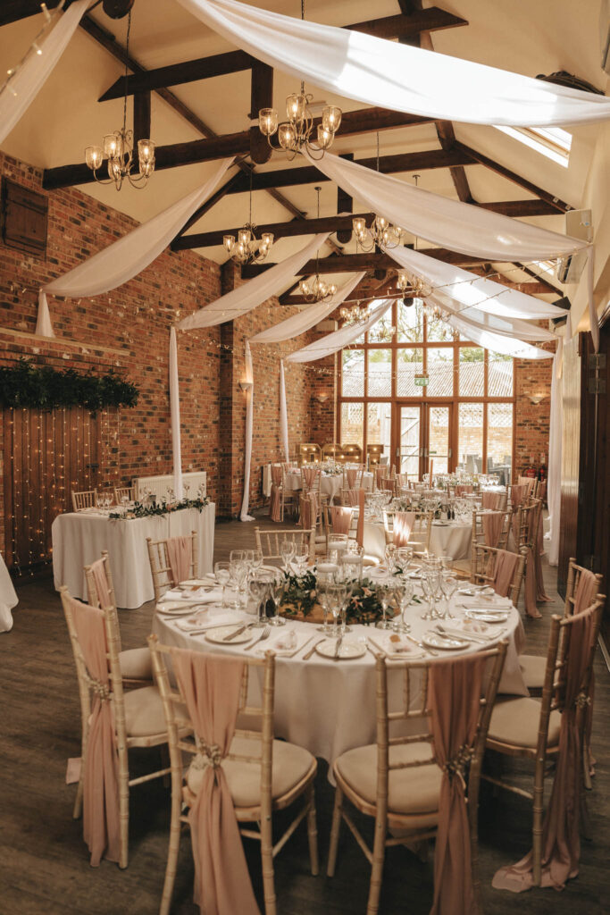 A rustic wedding reception hall in Waltham features round tables with white tablecloths and pink draped chairs. The ceiling is adorned with white fabric and chandeliers. Natural light pours in through large windows, and a backdrop of exposed brick walls adds warmth to the elegant setting. © Aimee Lince Photography