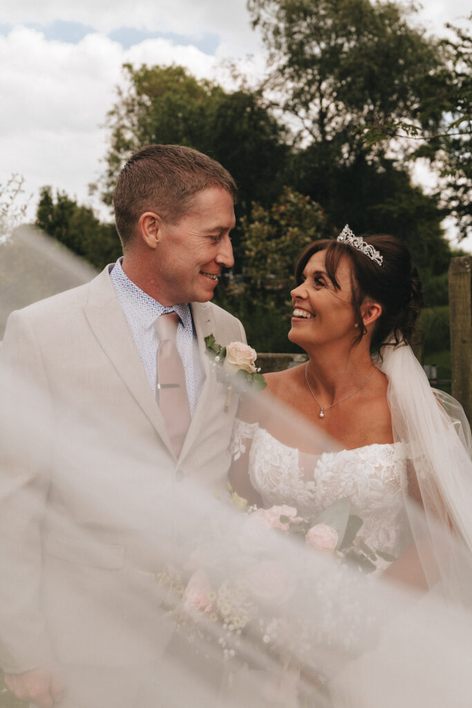 A bride and groom stand outdoors in Waltham, smiling at each other. The bride wears an off-shoulder white gown with a tiara, holding pink roses. The groom dons a beige suit and polka dot shirt. A sheer veil softly blurs the scene, set against green Lincolnshire trees in the background. © Aimee Lince Photography