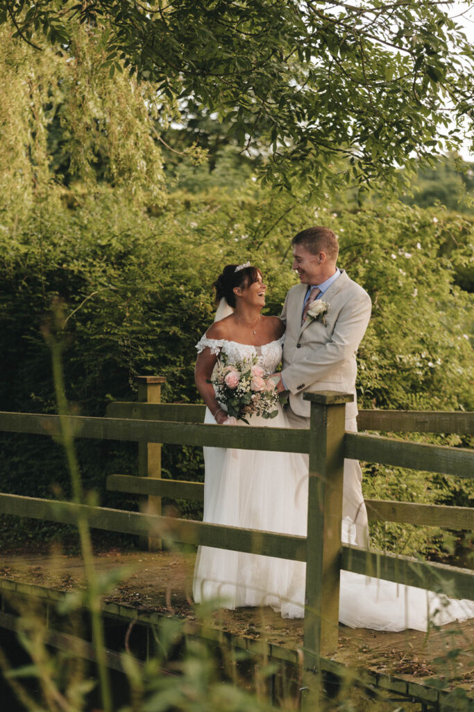 A bride and groom stand on a wooden bridge surrounded by the lush greenery of Lincolnshire. The bride wears an off-the-shoulder white gown, clutching a bouquet of pink and white flowers, while the groom dons a light suit with a boutonniere. They smile at each other under a canopy of trees. © Aimee Lince Photography