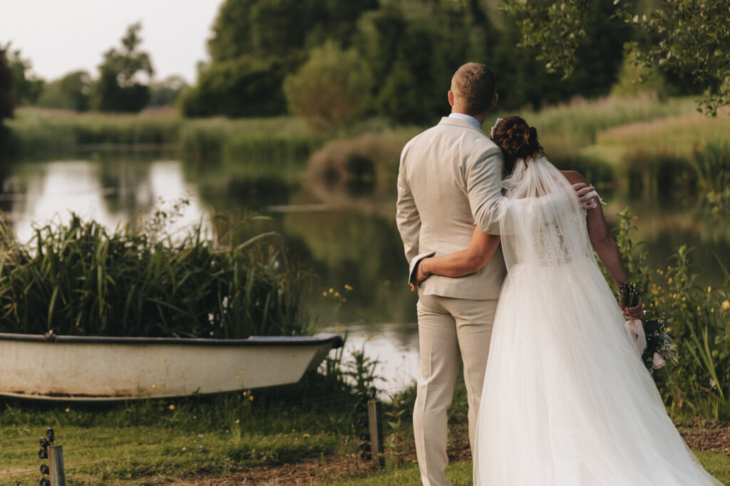 A couple stands by a serene lake at Hall Farm Hotel in Lincolnshire, embracing and gazing at the water. The bride wears a flowing white gown, while the groom is in a light beige suit. Nearby, a small rowboat rests amidst lush greenery under a clear sky, highlighting their magical wedding moment. © Aimee Lince Photography
