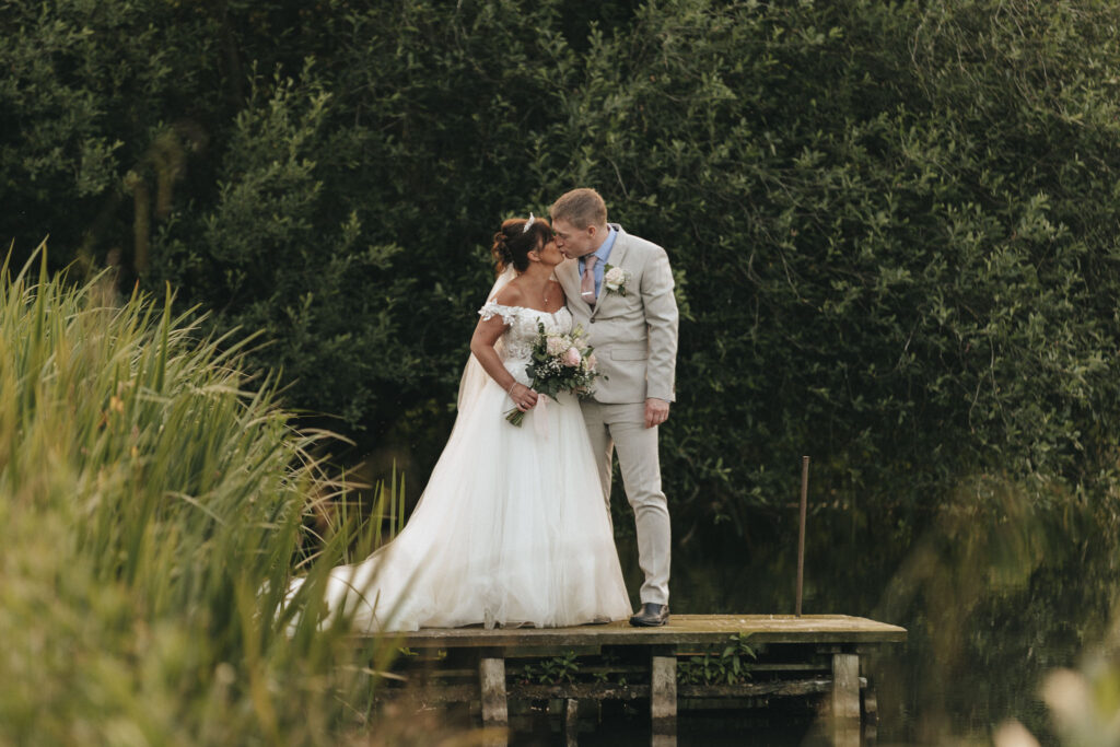 A bride in a white gown and a groom in a light gray suit stand on a wooden dock at Hall Farm Hotel, surrounded by lush greenery. The bride holds a bouquet as they share a kiss. The backdrop of dense foliage creates a serene, natural setting for their wedding by the water. © Aimee Lince Photography