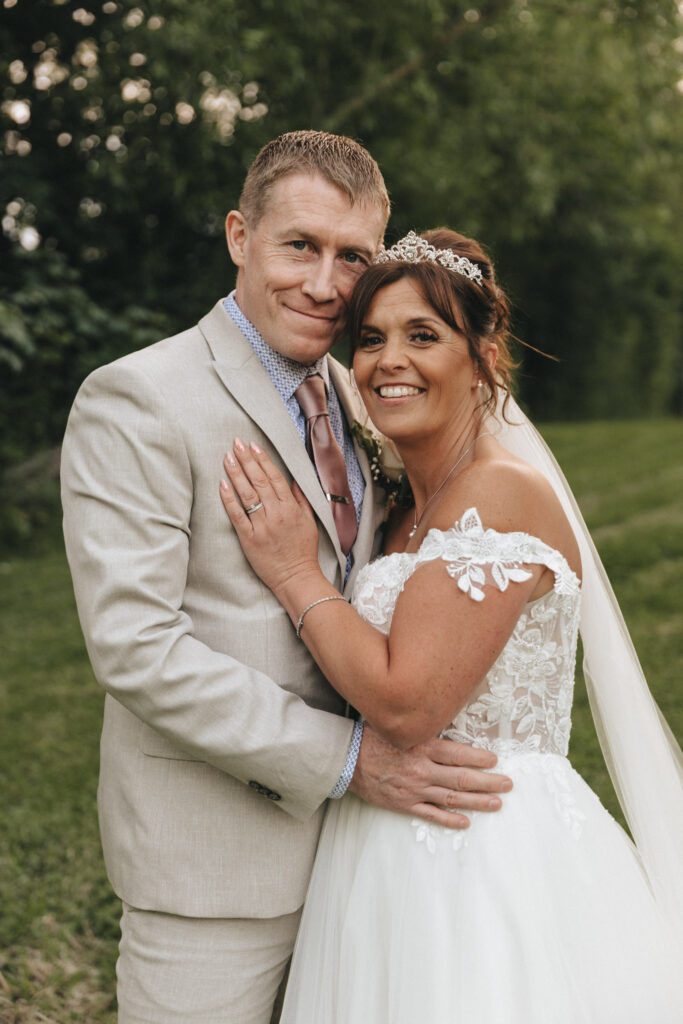 A couple dressed in wedding attire stands closely together outdoors at Hall Farm Hotel. The groom is in a beige suit with a patterned shirt and pink tie. The bride wears a white gown with lace, a veil, and a tiara. They smile warmly, surrounded by Lincolnshire's lush greenery. © Aimee Lince Photography