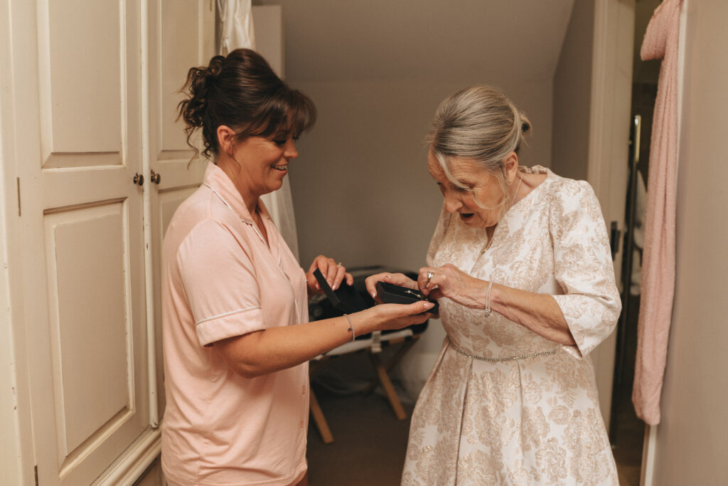 In a softly lit room with cream-colored cabinets, a woman in a pink outfit hands a small, dark box to another woman with gray hair and a patterned dress. Both appear joyful, the older woman looking surprised and excited, much like receiving delightful news at the Hall Farm Hotel in Lincolnshire. © Aimee Lince Photography