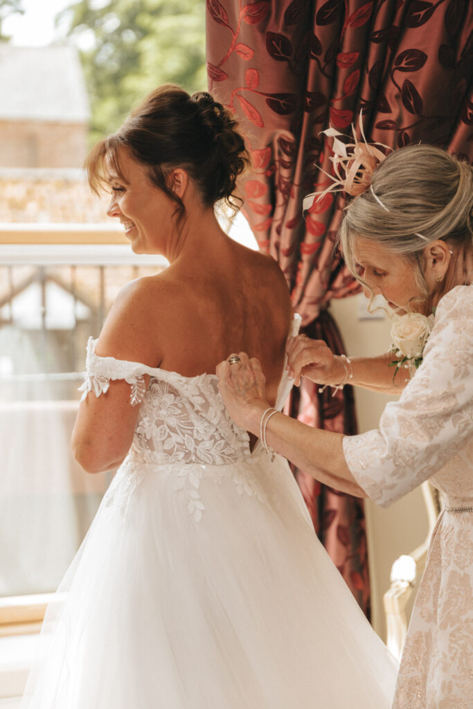 A bride in an off-shoulder lace wedding dress smiles as an older woman, perhaps her mother, helps fasten the gown by the window with red curtains. The scene unfolds in a charming Grimsby venue, where natural light filters through, enriched by the joy of wedding preparations. © Aimee Lince Photography