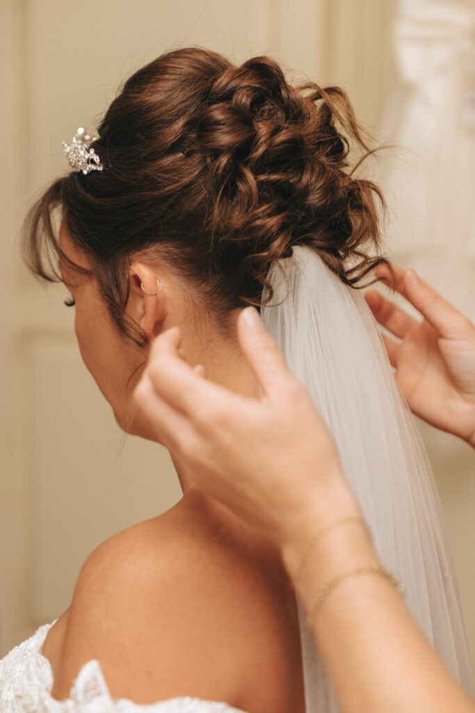 A person with wavy brown hair styled in an intricate updo, adorned with a sparkling tiara and a white veil, is being adjusted by another's hands. The setting suggests wedding preparations in Grimsby. The softly blurred background emphasizes the person preparing for the special day. © Aimee Lince Photography