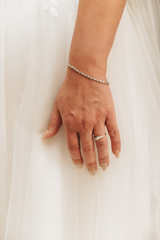 A close-up of a bride's hand wearing a sparkly bracelet and a solitaire diamond ring at her enchanting Lincolnshire wedding. Her fingers have a natural manicure, gently holding the flowing fabric of her white wedding dress, creating an elegant and delicate appearance. © Aimee Lince Photography