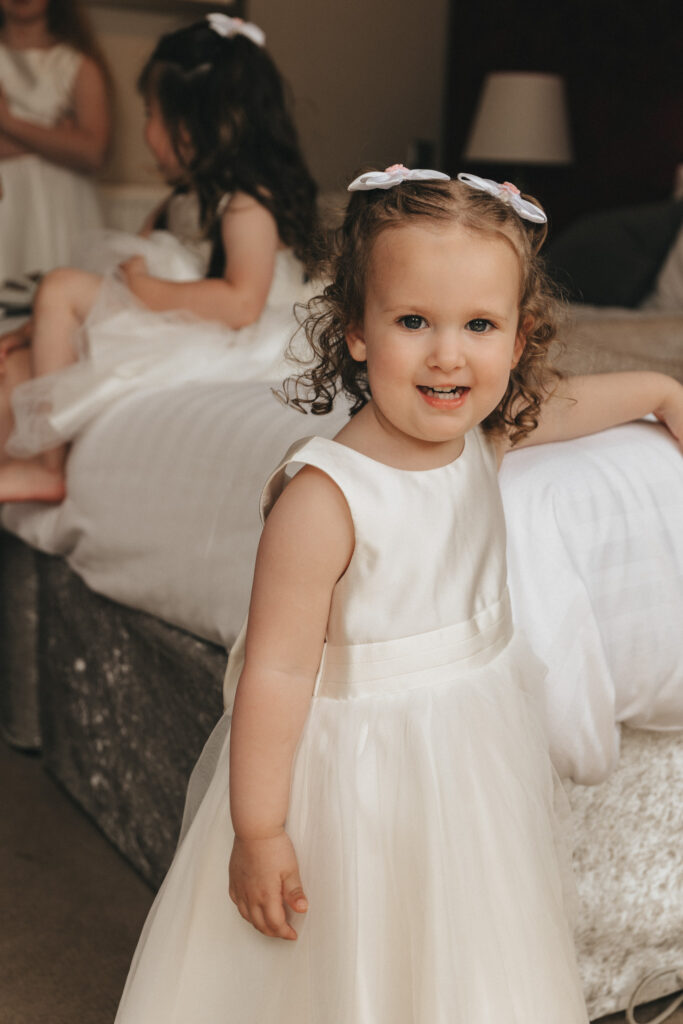 A young girl in a sleeveless white dress smiles at the camera. Her curly hair is adorned with a white bow. She stands beside a bed with another child in a similar dress visible in the background, reminiscent of cozy family gatherings in Waltham. The room has soft lighting, creating a warm and joyful atmosphere. © Aimee Lince Photography