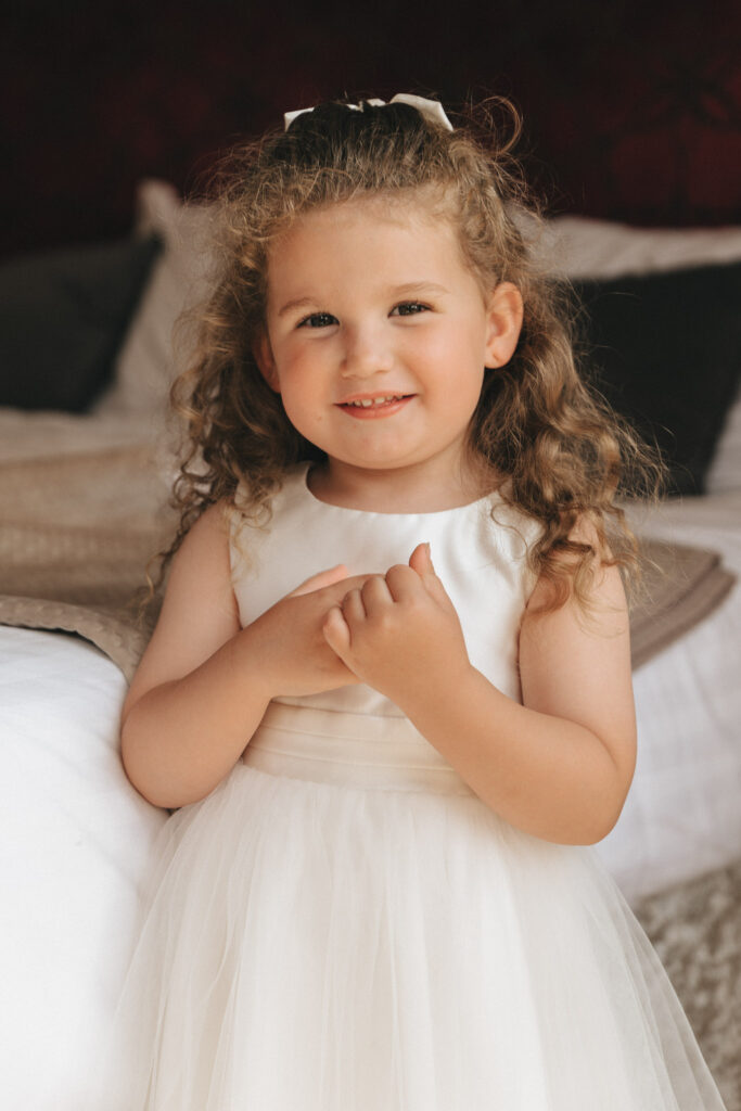 A young girl with curly brown hair and a white bow stands indoors at Hall Farm Hotel, smiling softly. She wears a sleeveless white dress, her hands gently clasped near her chest. The background features a neatly made bed with dark pillows, creating a warm and cozy atmosphere in Grimsby. © Aimee Lince Photography
