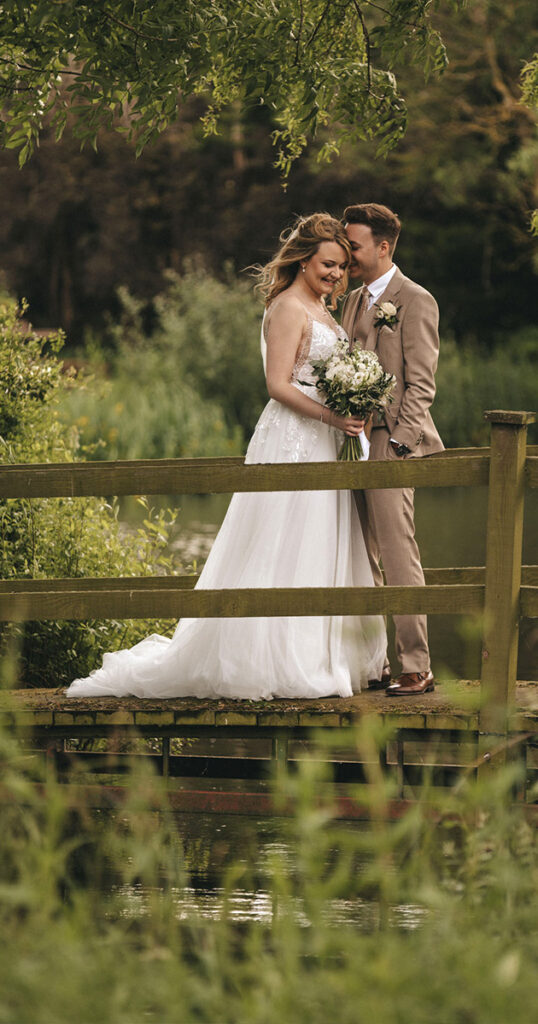 A bride in a white gown and a groom in a tan suit stand on a wooden bridge at Hall Farm Hotel, surrounded by lush greenery. The bride holds a bouquet, and they share smiles in tender harmony. The verdant setting near Grimsby adds to the serene, romantic backdrop with trees and water glimpses. © Aimee Lince Photography