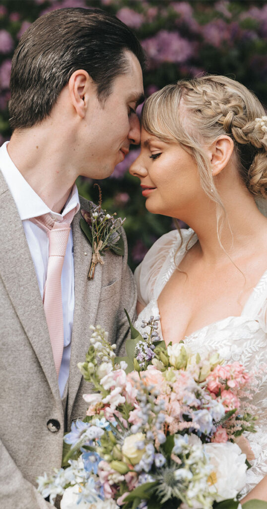 At Woodhall Spa Manor, a bride and groom gently touch foreheads, eyes closed in an intimate wedding moment. The groom wears a beige suit and floral boutonniere, while the bride in lace holds a colorful bouquet. Surrounded by purple flowers, the romantic ambiance is truly enchanting. © Aimee Lince Photography