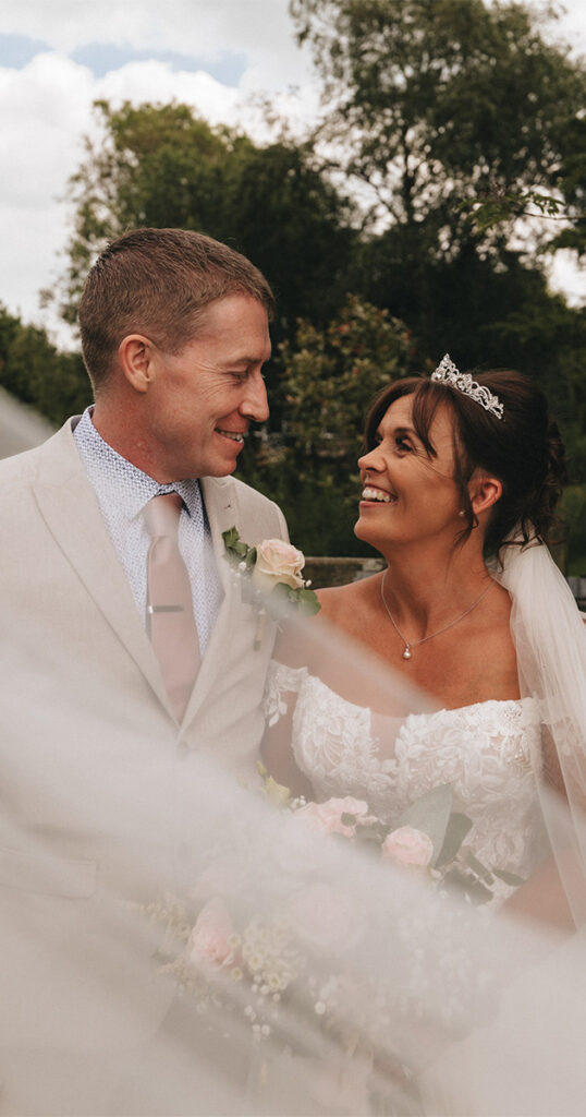 A bride and groom smile at each other outdoors at Hall Farm Hotel, creating a joyful and romantic atmosphere. The bride in a tiara, lace dress, and veil holds a bouquet. The groom wears a beige suit with a patterned shirt. Soft focus foreground and lush greenery enhance the serenity of this Grimsby setting. © Aimee Lince Photography