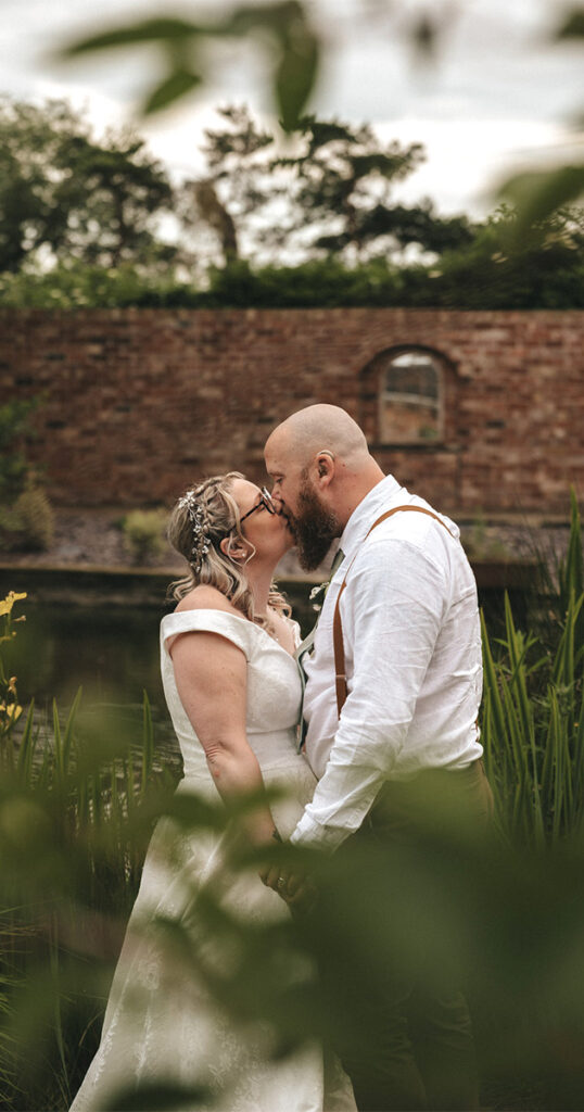 A couple kiss outdoors, framed by green foliage, capturing a serene wedding moment. The bride, in a white lace gown with a floral hair accessory, and the bearded groom in suspenders stand near a pond against a brick wall backdrop—an enchanting scene ideal for wedding photography. © Aimee Lince Photography