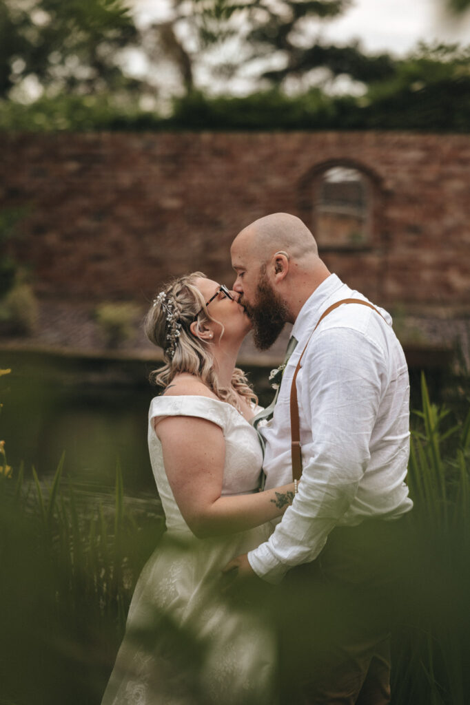 A couple shares a tender kiss outdoors against a brick wall and window at Stallingborough Grange Hotel. The woman, in a lace dress and floral headpiece, and the man, in a white shirt with suspenders, embody timeless wedding photography. Green foliage partially frames the intimate scene. © Aimee Lince Photography - Wedding photographer in Lincolnshire, Yorkshire & Nottinghamshire