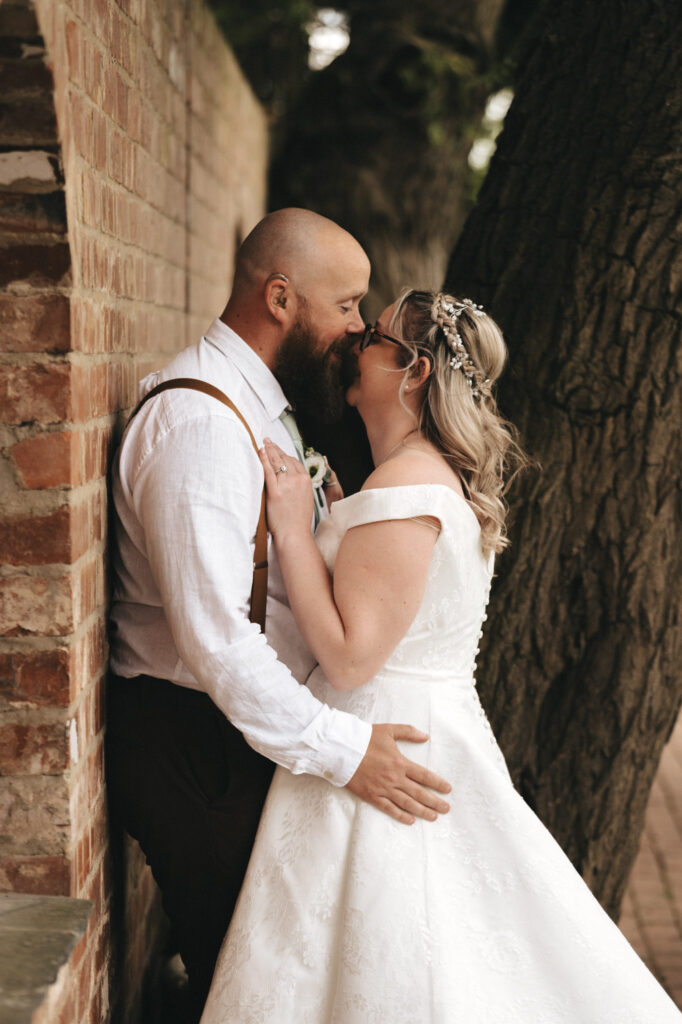 A couple stands closely under a tree beside a brick wall at Stallingborough Grange Hotel. The man wears a white shirt with suspenders, and the woman dons an off-the-shoulder white wedding dress with a floral hairpiece. They lovingly touch noses, sharing an intimate moment in the soft natural lighting. © Aimee Lince Photography