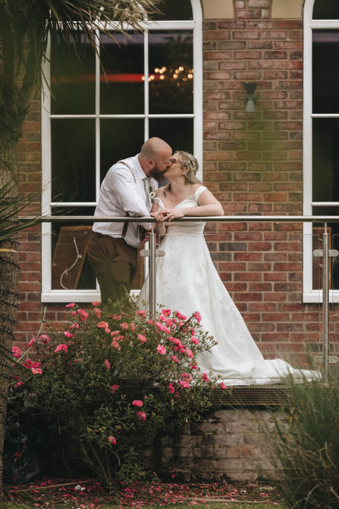 A newlywed couple shares a kiss on a balcony at Stallingborough Grange. The groom wears a white shirt and brown pants, while the bride is in a flowing white gown. They stand near a glass railing, with vibrant pink flowers and lush greenery below, set against a brick wall with arched windows. © Aimee Lince Photography