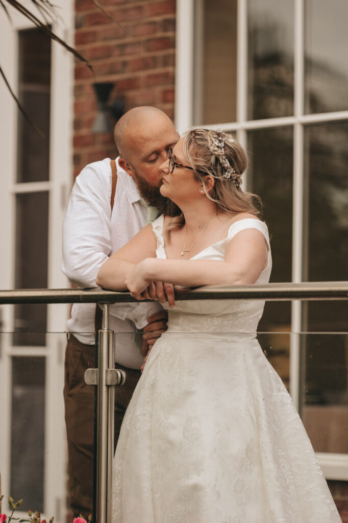 A couple embraces on a balcony at Stallingborough Grange. The man, with a beard and in a white shirt, gently kisses the woman on the forehead. Wearing glasses and a white wedding dress, she leans on the railing and smiles. A brick wall and glass doors are visible in the background. © Aimee Lince Photography