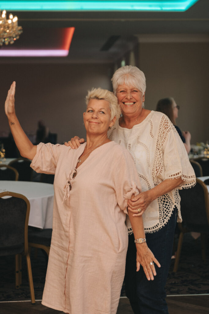 Two women smiling and posing in a warmly lit room at Stallingborough Grange. The woman on the left is raising one hand, wearing a light pink dress, while the woman on the right, in a white lace top, rests her arm on her friend's shoulder. Perfect for elegant wedding photography with chandelier charm above them. © Aimee Lince Photography