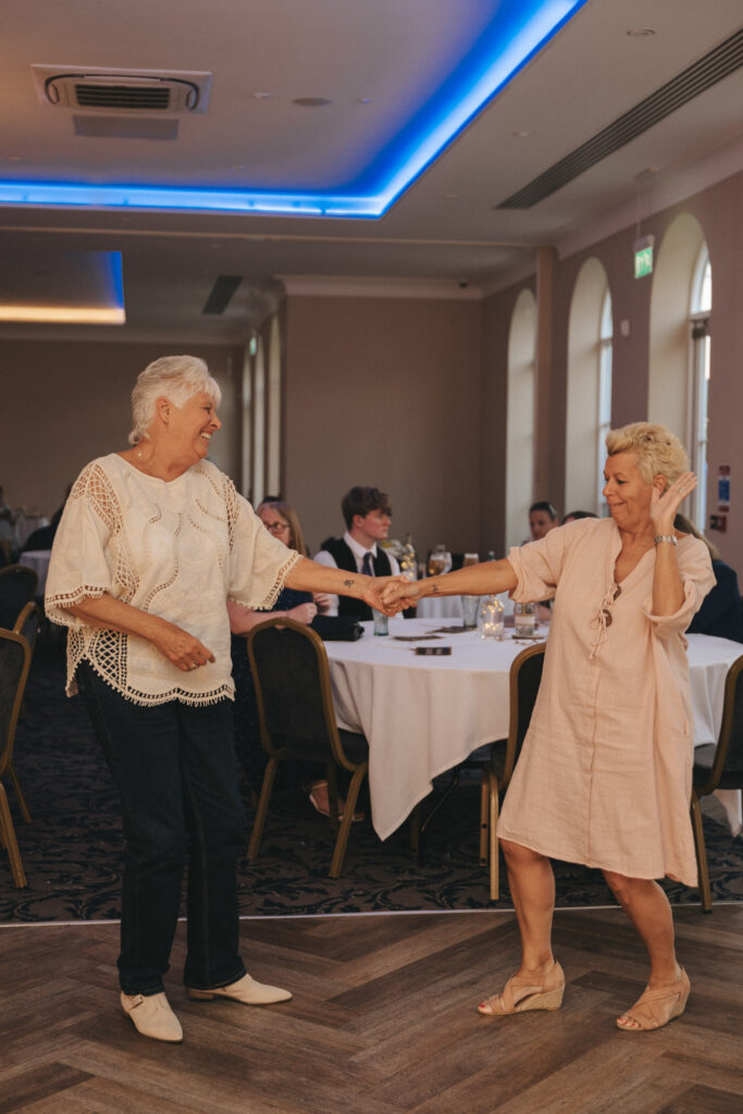 Two smiling older women dance joyfully in a warmly lit event space at the Stallingborough Grange Hotel. One wears a white top and jeans, while the other dons a light pink dress with a floral pattern. They hold hands, surrounded by round tables with white tablecloths, as guests watch their wedding celebration unfold. © Aimee Lince Photography