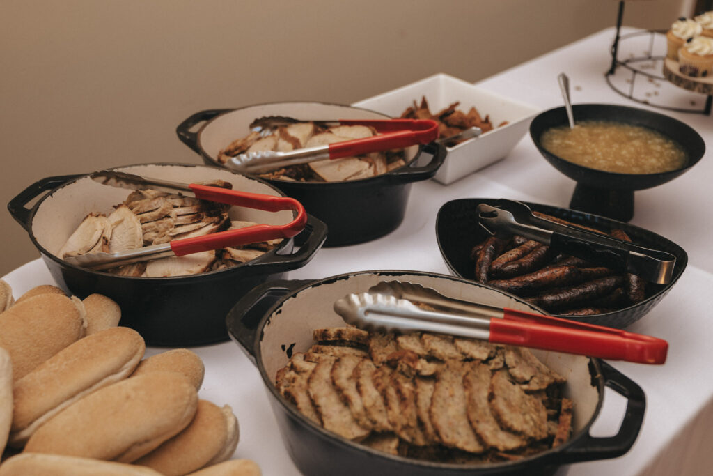 Amidst the elegant setting reminiscent of Stallingborough Grange Hotel, a table showcases sliced meats in black pans with red-handled tongs, sausages and hotdog buns, a bowl of thick sauce, and a white bowl with potato wedges—all neatly arranged on a white tablecloth. © Aimee Lince Photography