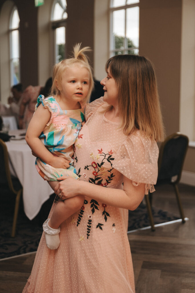 A woman in a pink, embroidered dress holds a young child in a colorful outfit at what appears to be a wedding. Indoors, with round tables and arched windows behind them, they share a tender moment together. This captivating scene beautifully captures the essence of wedding photography. © Aimee Lince Photography
