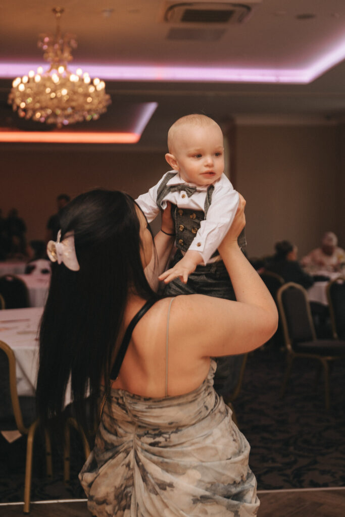 In an elegantly adorned room at Stallingborough Grange, a woman in a patterned dress holds a baby boy dressed in a crisp white shirt and vest. The enchanting scene, perfect for wedding photography, captures the baby's curious gaze while the woman's hair is tied with a large bow. © Aimee Lince Photography