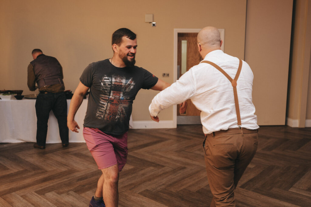 Two men dance joyfully indoors, perhaps caught in a candid moment of wedding photography. One wears a graphic T-shirt and pink shorts, while the other sports a white shirt with suspenders and brown pants. A person in the background sets up a table in this room with wood-patterned flooring and cream-colored walls. © Aimee Lince Photography