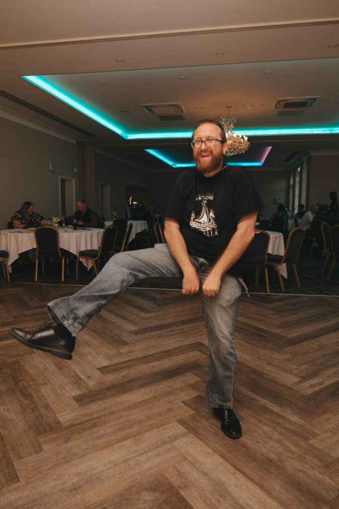 A bearded man with glasses and a black t-shirt, sporting jeans, is energetically dancing on the parquet floor of a dimly lit banquet hall at Stallingborough Grange Hotel. He lifts one leg while smiling, surrounded by elegantly set tables and guests, perfect for wedding photography beneath a grand chandelier. © Aimee Lince Photography