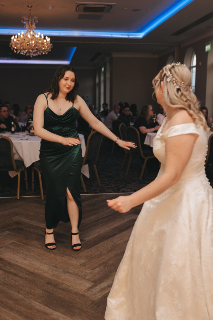 Two women are joyfully dancing at a wedding reception at Stallingborough Grange Hotel. The woman on the left wears a dark green dress, and the bride on the right wears a white gown. They twirl on a wooden dance floor under a chandelier, with guests seated at round tables beneath ambient blue lighting. © Aimee Lince Photography