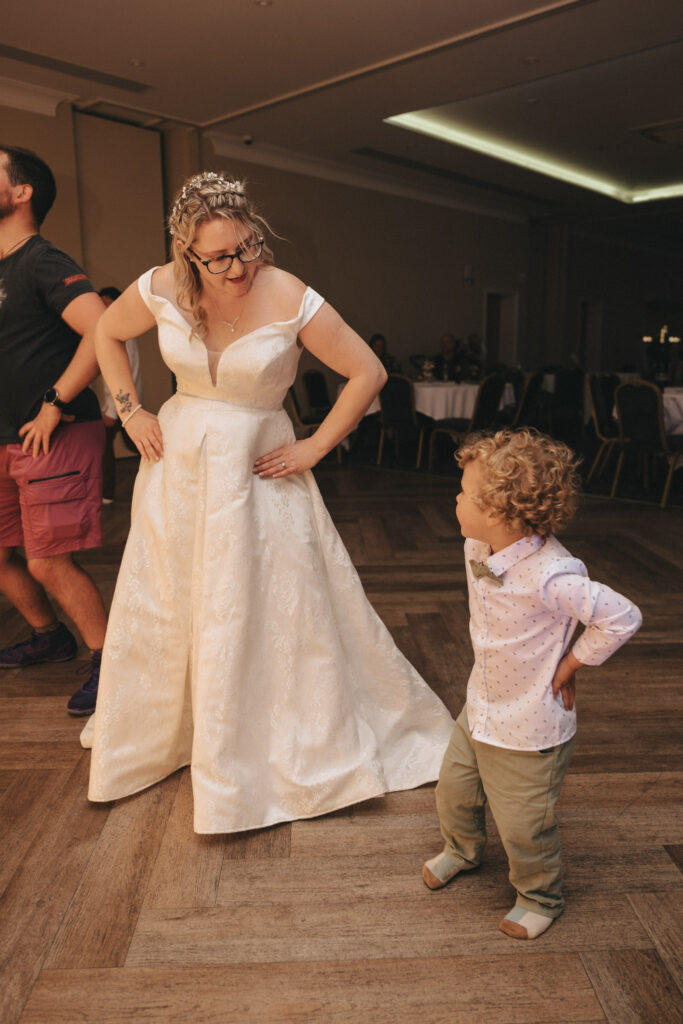 A woman in a white wedding dress and glasses stands confidently with her hands on her hips, smiling at a young curly-haired child in a white shirt and green pants. Captured at Stallingborough Grange Hotel, the room features wooden flooring, tables with chairs, and another guest partially visible on the left. © Aimee Lince Photography - Wedding photographer in Lincolnshire, Yorkshire & Nottinghamshire