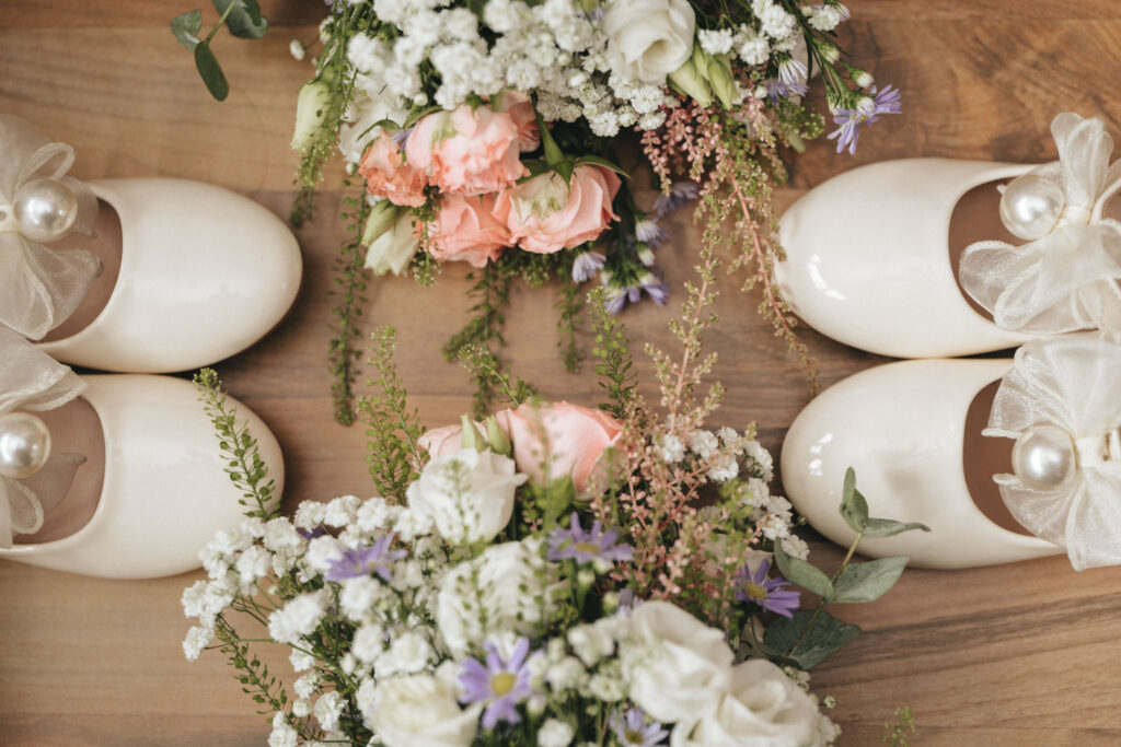 Stallingborough Grange Hotel Wedding Photographer - Steph & Kurt © Aimee Lince Photography (84) Close-up of four pairs of cream-colored flat shoes adorned with sheer bows and large pearls, arranged around bouquets. The elegant setup, perfect for a wedding at Stallingborough Grange Hotel, features white, pink, and lavender flowers with green foliage on a wooden surface. © Aimee Lince Photography