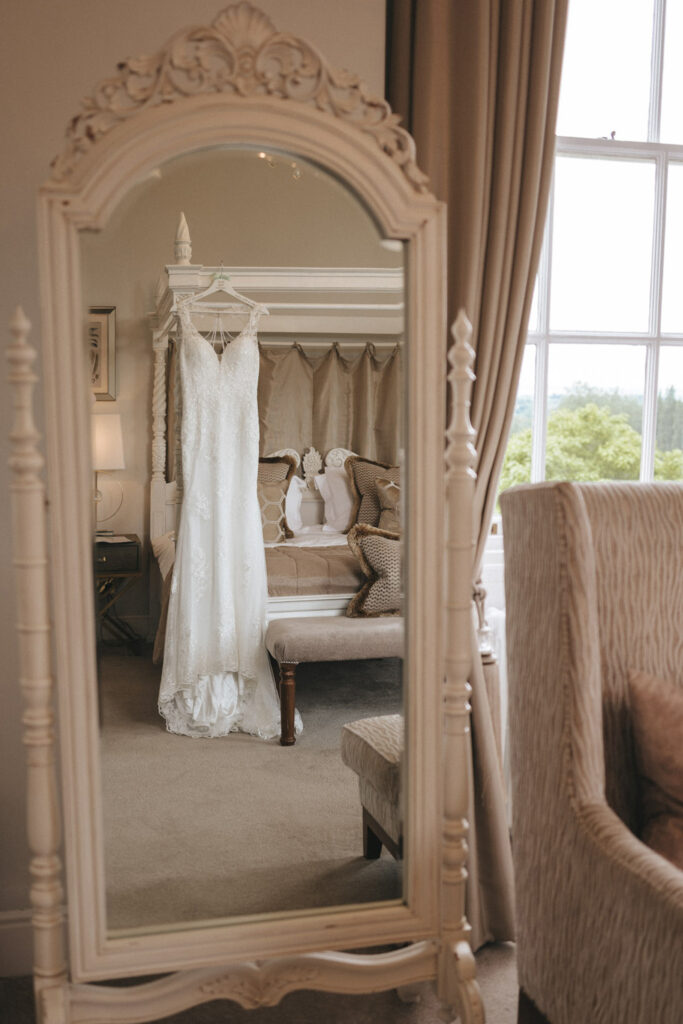 A white lace wedding dress hangs gracefully on a full-length antique mirror in a cozy, elegant room, perfect for capturing stunning wedding photos. The ambiance is warm and inviting at Bourton Hall Wedding venue, with soft natural light illuminating a plush armchair and neutral curtains. © Aimee Lince Photography