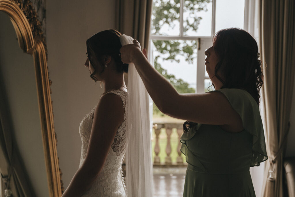 In a room at Bourton Hall Weddings, a bride stands beside a large mirror in her lace gown. Sunlight streams through the window, casting light on an individual in a green dress as they adjust the veil. A wedding photographer captures the moment against a backdrop of greenery and balcony railing. © Aimee Lince Photography