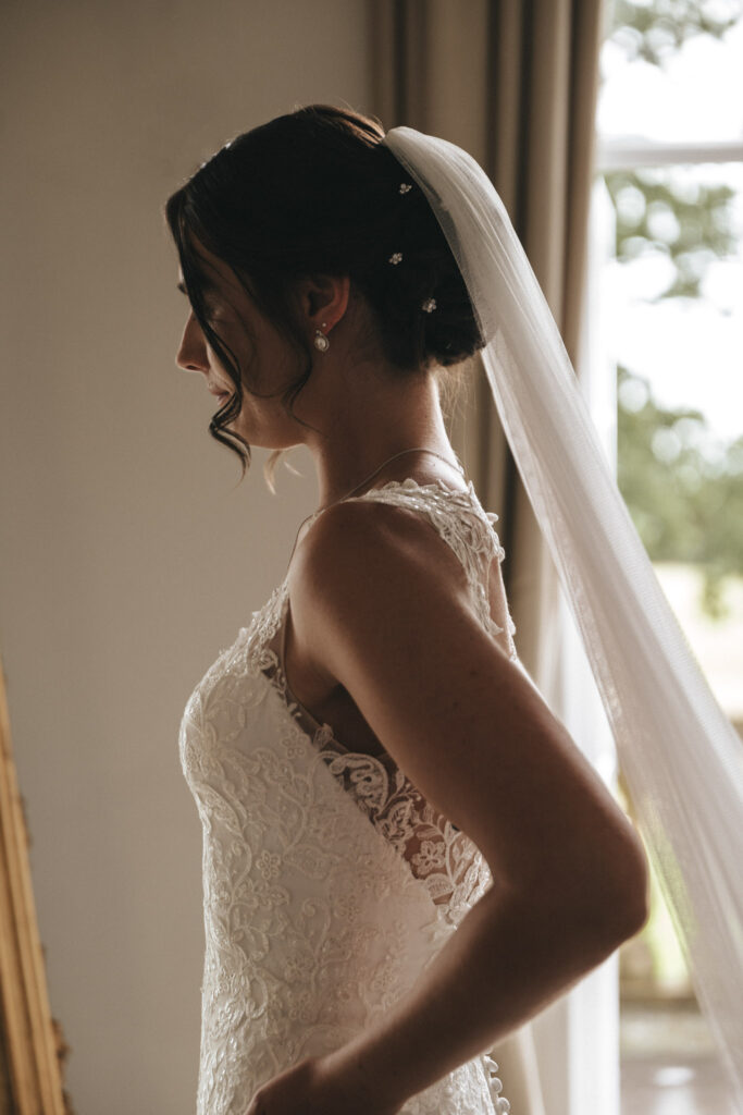 A bride in a lace wedding dress stands in a softly lit room at Bourton Hall. Her hair is styled in an elegant updo adorned with a long veil. She gazes out of a tall window, letting the natural light of Bourton Hall Weddings illuminate her profile, highlighting the intricate dress details. © Aimee Lince Photography