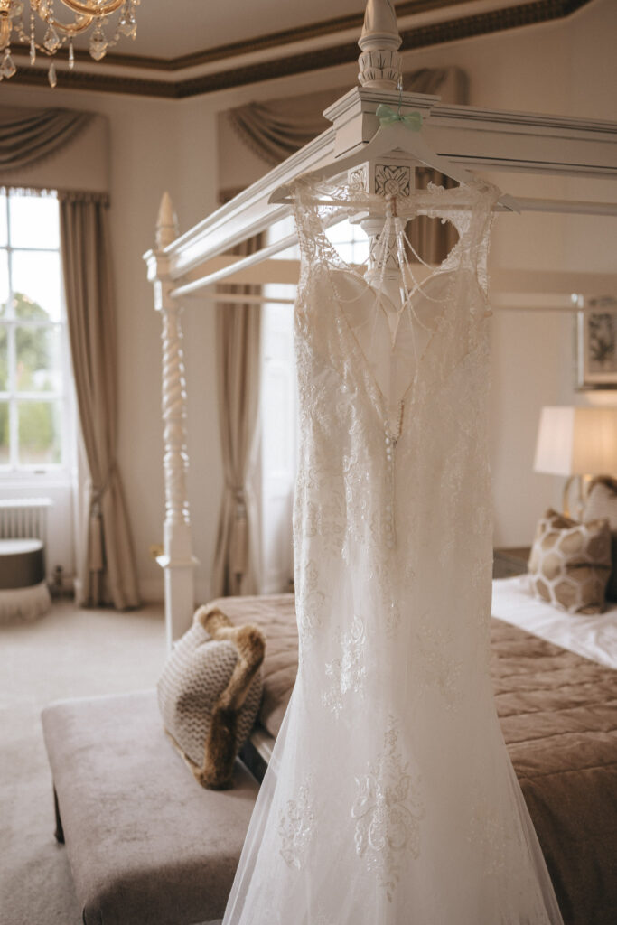 A delicate lace wedding dress hangs on a four-poster bed in an elegant room, perfect for wedding photography. The dress features intricate lacework and a sheer back. With beige curtains, a chandelier, and a plush bench with pillows, Bourton Hall Wedding photos capture a sophisticated and serene atmosphere. © Aimee Lince Photography