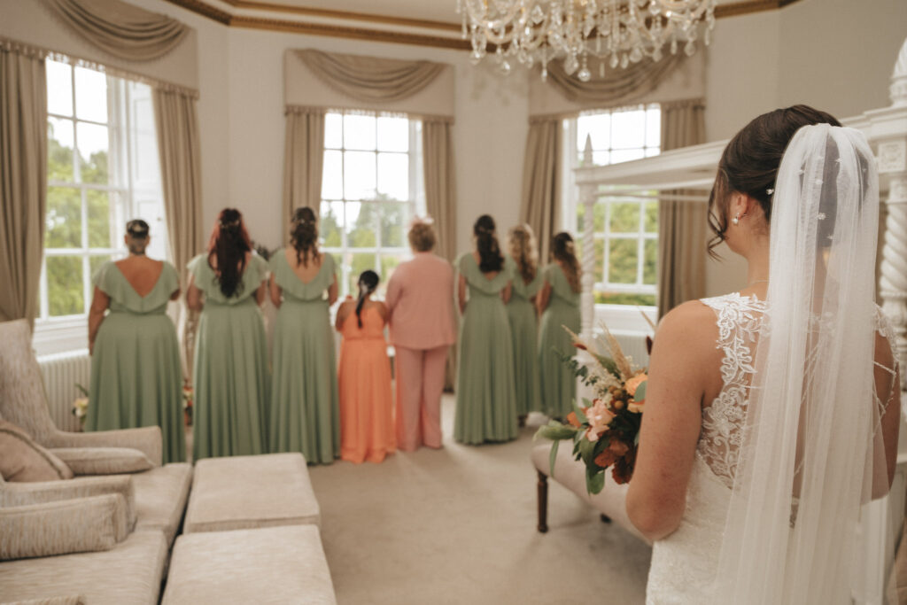 A bride in a white gown and veil holds a bouquet, facing a group against the elegant backdrop of Bourton Hall. The bridesmaids in green dresses, a young girl, and someone in pink attire pose for wedding photos in the bright room with large windows, neutral décor, and a chandelier. © Aimee Lince Photography