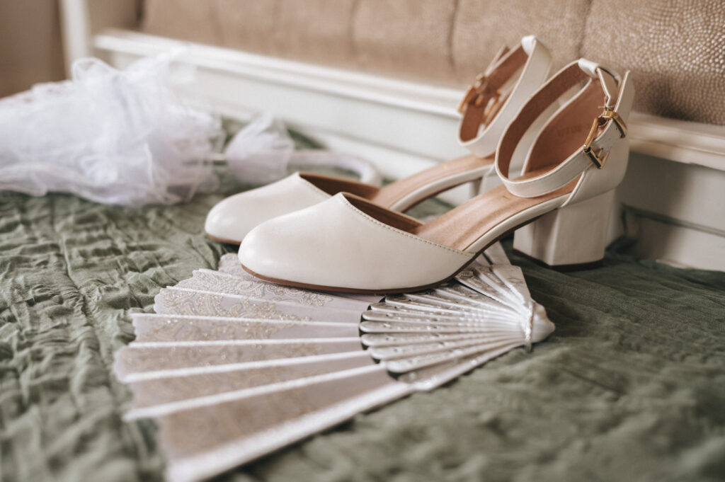 A pair of elegant, cream-colored block-heeled shoes with ankle straps rest on a textured green bedspread. Next to them, an ornate lace fan adds vintage charm. In the background, a white veil softly blurs, perfect for capturing timeless wedding photos at a Bourton Hall Weddings event. © Aimee Lince Photography