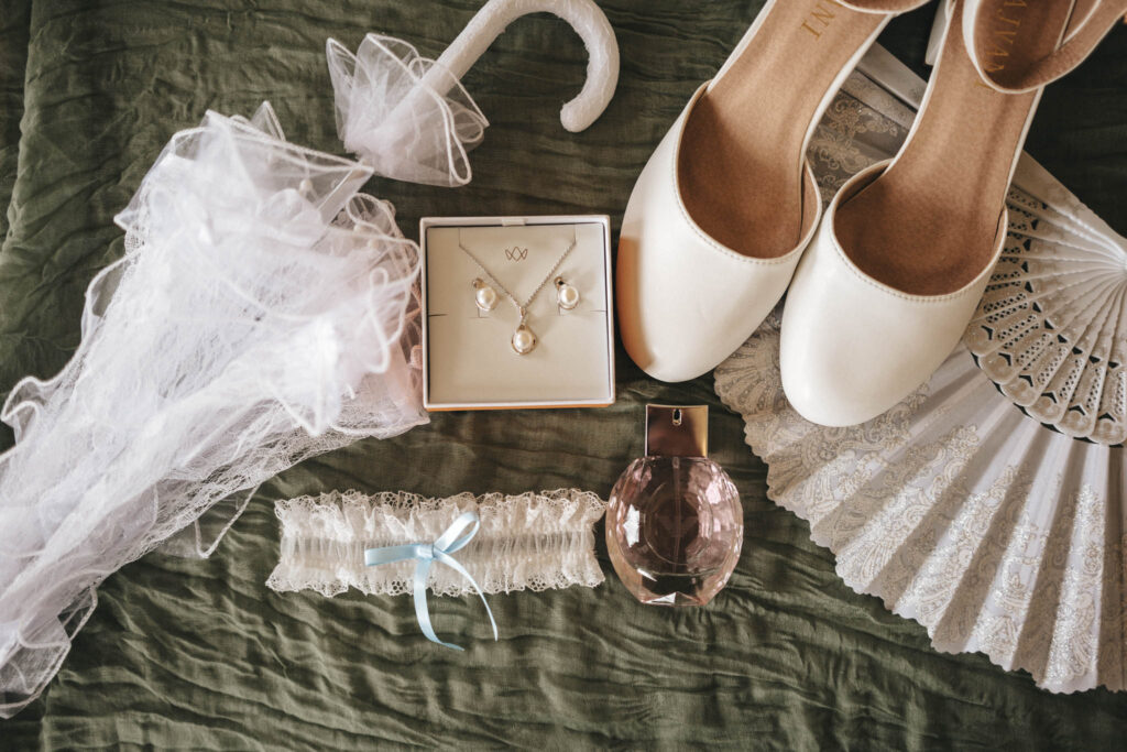 A flat lay of bridal accessories on a dark green fabric, perfect for wedding photography. Includes white heels, a lace garter with a blue ribbon, a perfume bottle, a jewelry set with earrings and necklace in a box, a lace fan, and a veil—ideal details for capturing memorable wedding photos. © Aimee Lince Photography