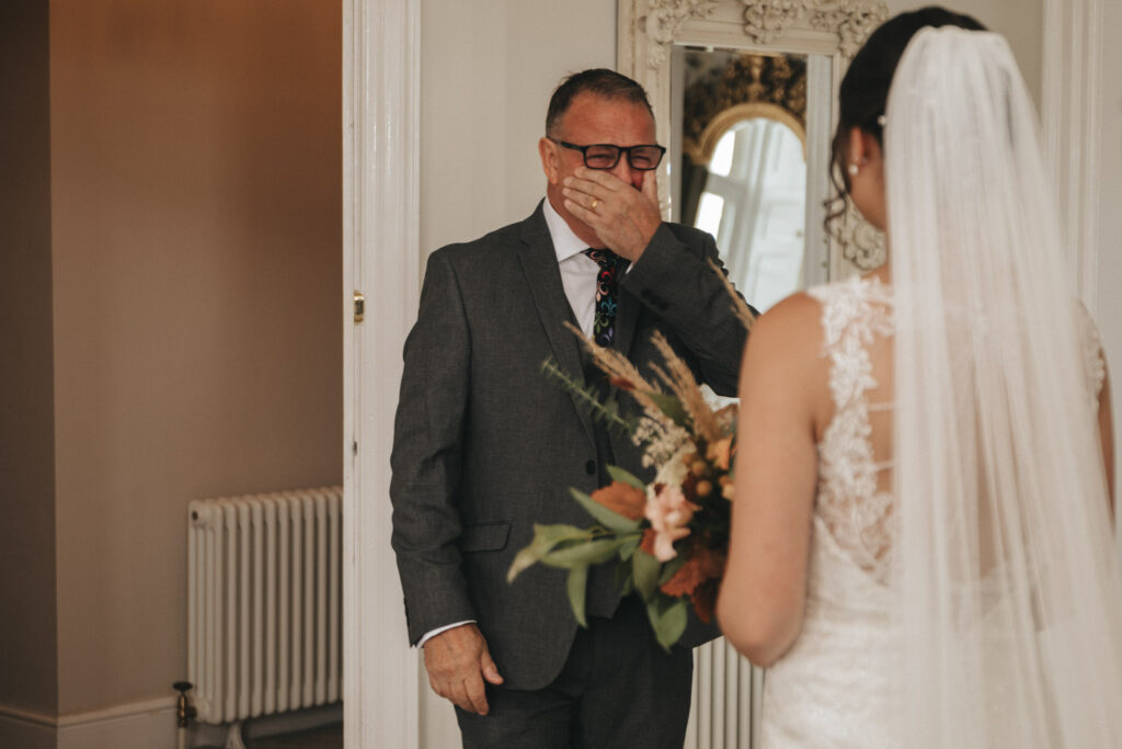 In a warmly lit room at Bourton Hall, a bride in a white lace dress holds a bouquet, facing her emotional groom in a gray suit. He wears glasses and a patterned tie. The decorative mirror and radiator add charm to their perfect wedding moment captured in timeless photos. © Aimee Lince Photography