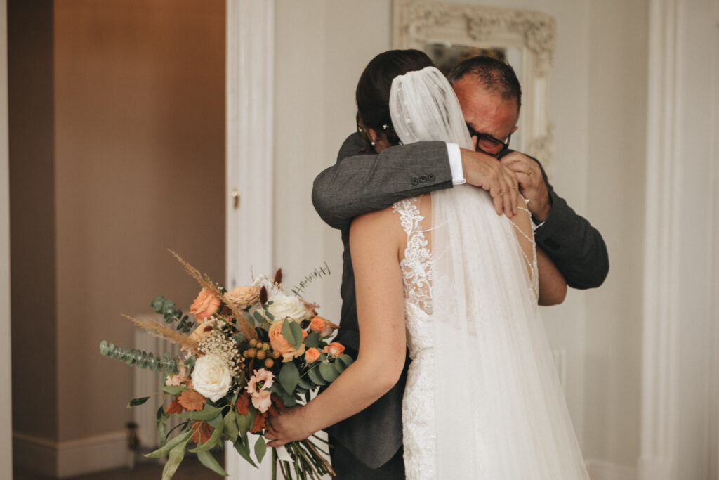 Bourton Hall Rugby Wedding Photographer - Olly & Jordan © Aimee Lince Photography (158) In the warmly lit room of Bourton Hall, a bride in a white lace gown embraces an older man in a gray suit, likely her father. She holds a bouquet of white roses and peach flowers. His eyes behind glasses reveal emotion. The moment is beautifully captured in their wedding photography. © Aimee Lince Photography