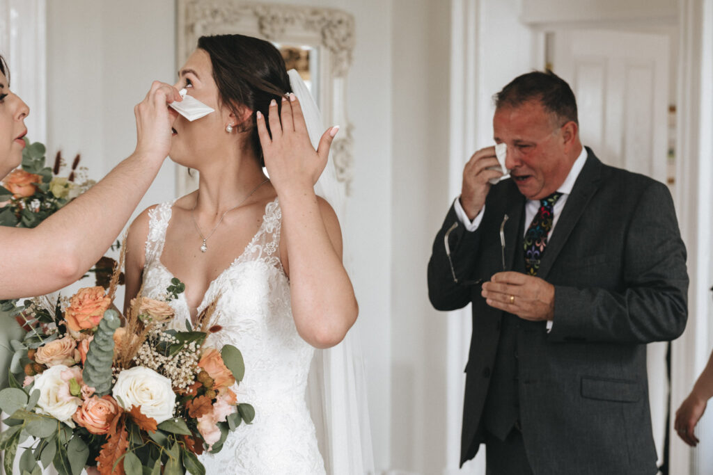 Emotions run high as a wedding photographer captures a bride in a white lace gown, wiping her tears with a tissue at Bourton Hall. Holding peach and white flowers, she stands beside another woman, who offers comfort. In the background, the lens catches a man in a suit also dabbing his eyes with emotion. © Aimee Lince Photography