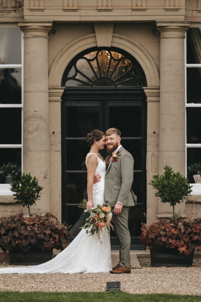 A bride and groom stand lovingly in front of an arched doorway, captured beautifully by a wedding photographer. The bride wears a white dress, holding a bouquet, while the groom dons a gray suit. Surrounded by potted plants against a stone building backdrop, the scene exudes classic elegance. © Aimee Lince Photography